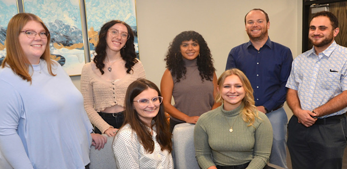 Group of seven GRILL lab team standing in a rown behind some that are sitting in a room smiling