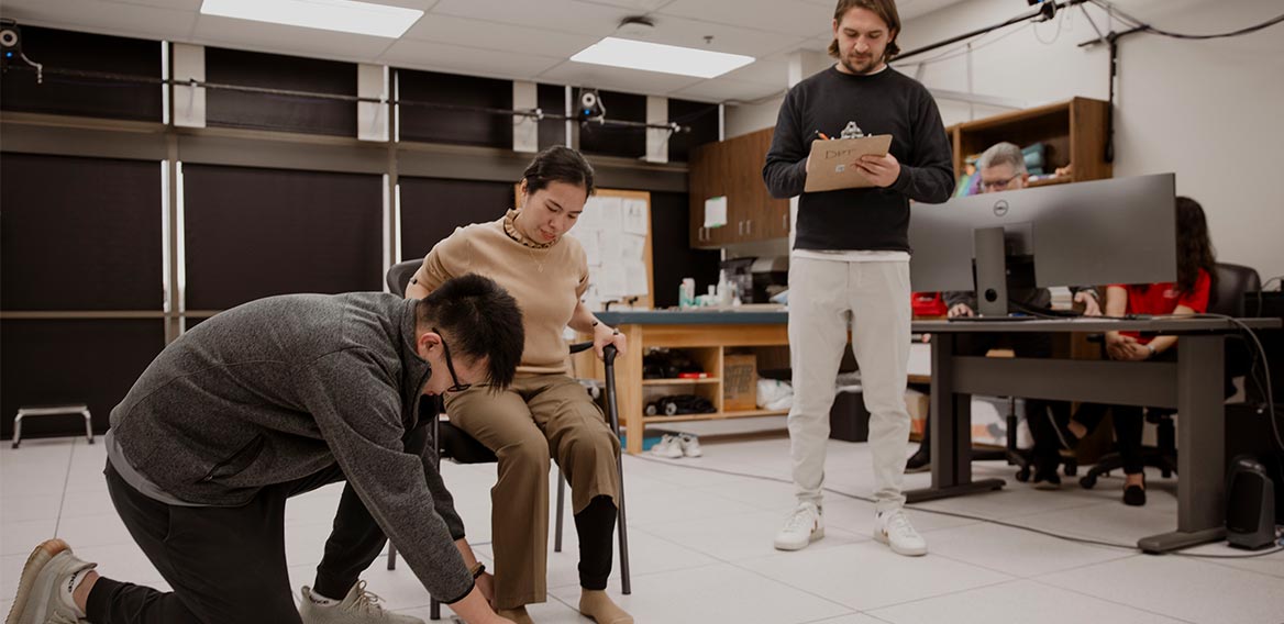 A group of students in a laboratory, with one sitting at a workbench and the others standing, collaborating on a research project