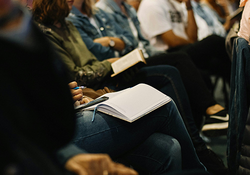 A row of people sitting with notebooks on top of their laps looking down wtih pencils in their hands