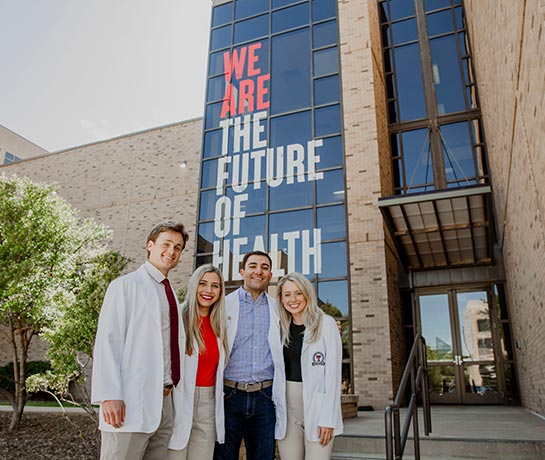 Four students standing infront of a building with we are the future of health sign behind them