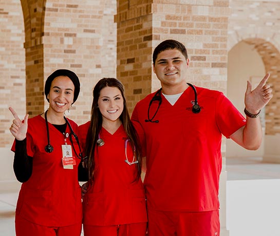 Three students in red scrubs standing next to each other smiling with tech guns up