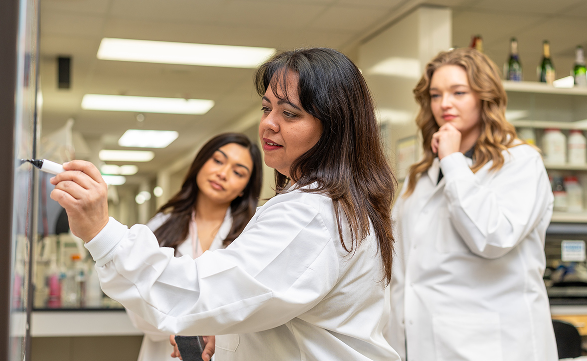 Female scientist in a white lab coat writing on a whiteboard while two graduate students observe in a laboratory setting.