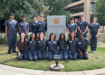 A group of TTUHSC Premedical Enrichment students in grey scrubs standing around the Anatomical Sciences Monument on the TTUHSC Lubbock campus outside
