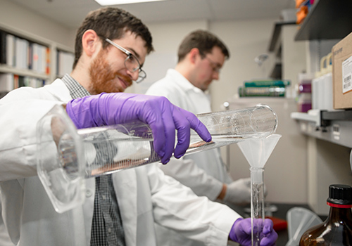 two TTUHSC students in white labcoats with rubber gloves on handling beakers pouring fluid into them