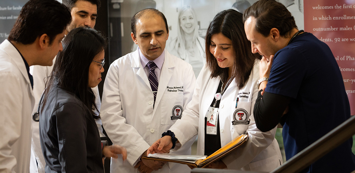 TTUHSC learners in white lab coats holding a binder standing in a circle on the Lubbock campus infront of the stair case and graphic wall looking down and smiling