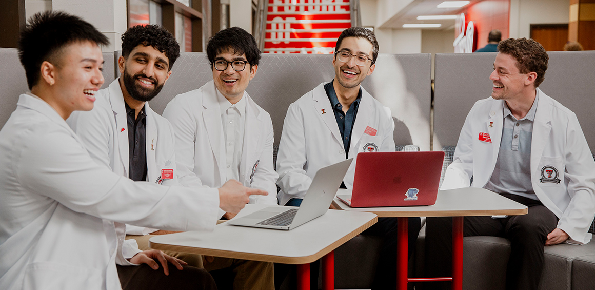Five TTUHSC students in white lab coats sitting at a table with computers smiling at each other