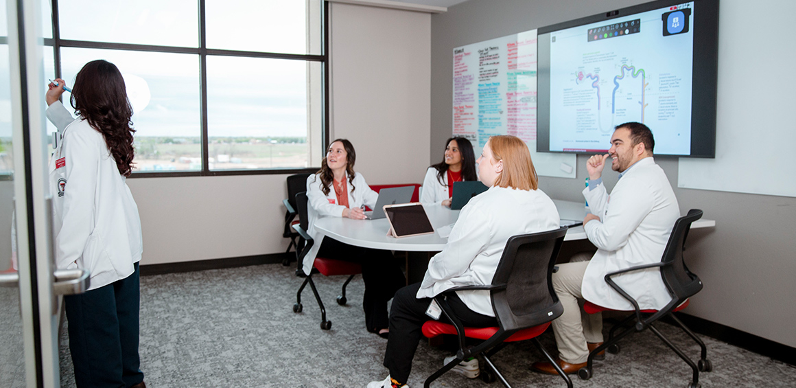 TTUHSC students in library study room in white lab coats sitting and smiling looking at another TTUHSC student using the dry erase board