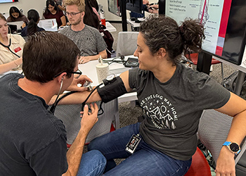Students sitting at a table while one student uses a blood pressure cuff on another students arm