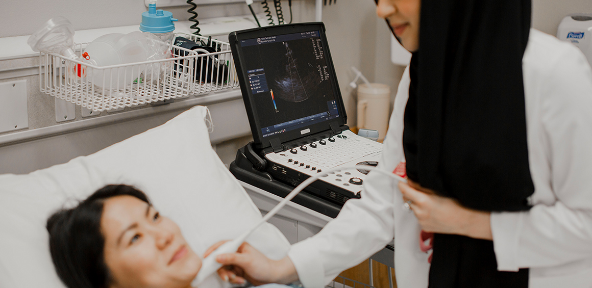 TTUHSC student in white lab coat standing over a patient in a bed holding a ultrasound transducer on the patients throat.