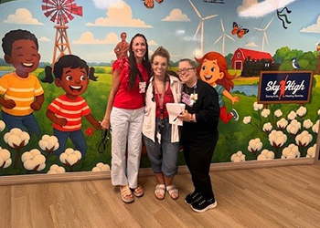 Three women smiling in front of a colorful mural featuring children, windmills, and cotton fields with a sign that reads “Sky High."