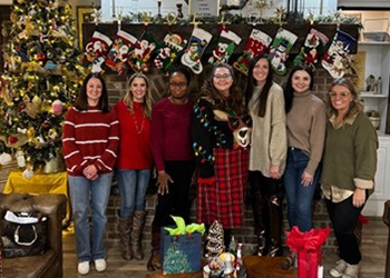 Seven women standing in front of a decorated fireplace with stockings and a Christmas tree, celebrating the holidays.