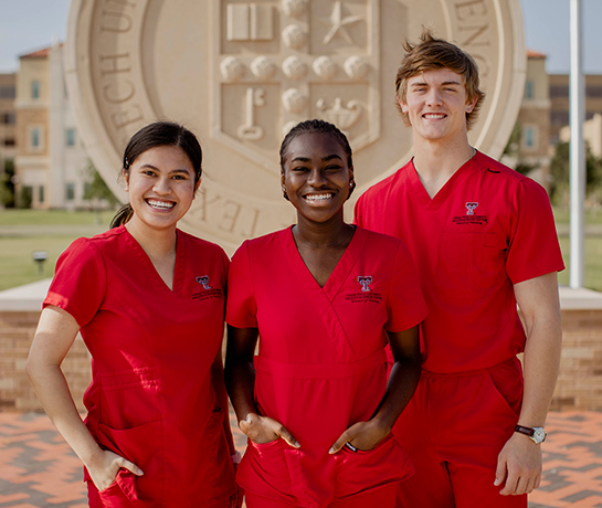 three TTUHSC nursing students in red scrubs smiling standing infront of the TTUHSC seal outside on the Lubbock campus